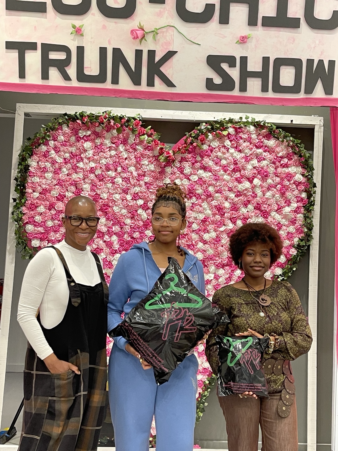 Dr. Bonita Best with two young women holding Runway Recycled garment bags at the Eco-Chic Trunk Show, in front of a pink and white rose heart wall